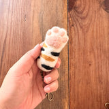 A female hand gently holds a handmade felt cat paw keychain against a warm wooden backdrop. This unique felt keychain features a white paw pad with textured pink toe beans and black and brown stripes around the wrist. A copper keychain with a clasp is attached.