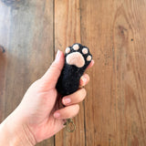 A black felt cat paw keychain is gently held by a female hand against a rustic wooden backdrop. This handmade felt keychain features a realistic cat paw design with puffy pink paw pad details. A copper keychain with a clasp is attached, completing the unique design.