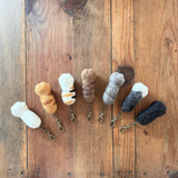 Seven unique felt cat paw keychains are displayed in a row on a rustic, weathered wooden surface, evoking warmth and homespun charm. Each handcrafted felt keychain is a detailed 1:1 replica of a cat paw, featuring distinctive paw pads and finger details. They come in various colors and patterns, including white, orange striped, white with black stripes, and shades of gray and black, each attached to a metal clasp.