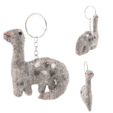 A handmade brontosaurus felt keychain is showcased against a bright white background. The soft, mottled gray wool felt brontosaurus features a long neck, rounded body, small tail, black eyes, and a red smile, accented with white and dark gray felt spots. This unique dinosaur keychain is attached to a metal keychain with a key ring.