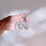 A gray wool felt Brontosaurus dinosaur keychain is gently held by a female hand against a soft white background with subtle light patterns. Crafted from textured gray wool felt, this handmade dinosaur keychain features darker spots along its body and a small red detail for the mouth. A metal keychain is attached, completing its unique, handcrafted look.