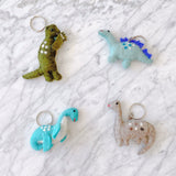 Multiple handmade wool felt brontosaurus keychains are arranged on a bright white marble backdrop. These unique keychains feature slight variations in texture, including a moss green brontosaurus with white spots, light blue, and gray designs. Each charming dinosaur figure is equipped with a functional metal keychain and key ring.
