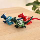 Three wool felt flying dragon keychains are displayed in a row on a light wooden surface, with a soft green leafy plant in the blurred background. Each handmade dragon keychain features tiny legs, horns, and tails, with wings accented by contrasting white details. The keychains come in blue, green, and red, with the green dragon in the foreground showcasing a copper clasp, creating a whimsical and charming scene.