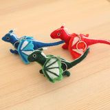 Three handcrafted felted wool flying dragon keychains are displayed on a wooden surface. A blue dragon keychain with a light blue and white wing is at the back, next to a red dragon keychain featuring a red and white wing. In front, a green dragon keychain has a light green and white wing. Each charming dragon keychain includes a small horn, tail, tiny feet, and a copper clasp.