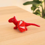 A vibrant red wool felt flying dragon keychain rests on a light brown wooden surface, with a blurred green plant in the background. This handcrafted felt keychain features white horns, white-accented wings, and black eyes. A metal clasp is attached to its back.