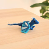 A handcrafted wool felt flying dragon keychain is displayed on a light wooden surface, set against a bright, airy background with blurred greenery. The unique blue dragon features a darker blue body with lighter blue and white accents on its wings and tail, and small white horns. A metal keychain with a clasp is attached to its back.