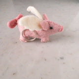 A playful wool felt flying pig keychain rests on a light marble surface in a close-up studio shot. This pink handcrafted keychain features white wool felt wings, pink ears, a round snout, and a curly tail, creating an endearing and whimsical appearance. A metal keychain is attached to its body.