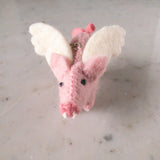 A handcrafted wool felt flying pig keychain rests on a light, neutral surface. This whimsical light pink pig features fluffy white wings, small perky ears, tiny black eyes, and a cheerful expression. A metal ring attaches to the top.