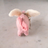 A handcrafted wool felt flying pig keychain is presented against a plain, light surface. This whimsical felt keychain features a plump, pink wool felt pig body with a curly tail and fluffy white wings attached to its back. A metal keychain with a clasp is connected to the top, highlighting its charming details.