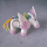 A charming wool felt pastel unicorn keychain rests on a mottled gray surface, highlighting its whimsical design. This handmade keychain features a white body with pink hooves and snout, and a playful mane with pastel yellow, green, purple, and pink strands. A silver-wrapped horn and a pink flower accent its side, completed with a metal key ring for attachment.