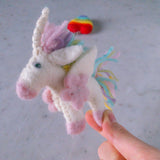 A handcrafted felt pastel unicorn keychain is held by a female hand against a light gray, mottled surface, with a blurred rainbow heart in the background. This whimsical unicorn is predominantly white with a pink snout, pink hooves, and light pink wings. Its mane is pastel pink and white, featuring a twisted silver horn and a colorful tail with teal, yellow, and pink strands.