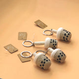 Three handcrafted felt Taiwanese bubble tea keychains are displayed on a plain peach surface, each with an attached small illustrated tag. These unique keychains are meticulously shaped like bubble tea cups from plush felt, featuring a light beige body with black speckles for tapioca pearls, a fluffy white lid, and a slender felt straw, all with a metal keychain.