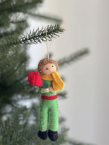 A whimsical felt Little Prince ornament, handcrafted from felted wool, hangs from a Christmas tree branch. This charming Christmas tree ornament features the Little Prince with brown curly hair, a smiling face, a green suit with a red belt, black shoes, and a yellow scarf, holding a red rose. A jute string allows it to hang gracefully against a soft, blurred white background.