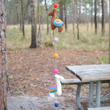 A wool felt Llama garland hangs playfully outdoors in front of a wooded area and a weathered picnic table. This handcrafted garland features two visible 3D felt llamas, one brown and one white, both adorned with colorful saddle blankets. Small, round felt balls in orange, pink, purple, and blue are interspersed along the string, adding a whimsical touch.