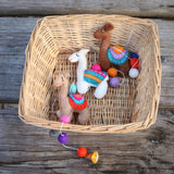 A colorful felt Llama Garland rests inside a woven basket on a rustic wooden surface, highlighting its handmade charm. The garland features three distinct three-dimensional wool felt llamas in white, brown, and tan, each adorned with unique colorful saddle blankets and decorative stitching. Vibrant felt balls extend from the garland, adding to its playful warmth.