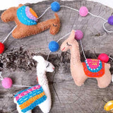The felt llama garland is displayed on a rustic wooden surface, creating a whimsical scene. This handcrafted wool felt garland features three distinct llamas interspersed with colorful felt balls. One llama is light brown with an orange, blue, and purple saddle; another is light pink with a red, pink, and blue saddle; and the third is white with a pink, blue, and orange saddle. Red, pink, purple, and blue felt balls complete the vibrant design.
