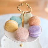 A set of handmade felt macarons is charmingly displayed on a decorative, tiered serving stand. These wool felt macarons feature a textured surface and white felt filling, mimicking real desserts in soft pastel shades of yellow, teal, green, peach, pink, and purple. Handcrafted with azo-free dyes for imaginative play, these fair-trade felt treats add sweetness and creativity to any setting.