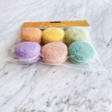 A package of six delightful felt macarons is presented on a light marble surface. These soft, round felt play food desserts, handcrafted from 100% wool with eco-friendly dyes, come in pastel shades of peach, yellow, light green, lavender, light pink, and turquoise. Each macaron features two colored felt pieces with a thin white layer, offering a charming and tactile design.