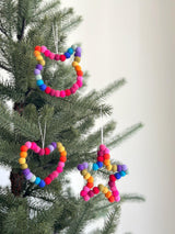 A vibrant Felt Mini Pompom Love Wreath Ornament is prominently displayed, hanging on a lush green Christmas tree. These 4-inch diameter ornaments are made from colorful 100% wool felt mini pom poms arranged in a joyful way. Suspended by a delicate jute thread, each adds a festive and handmade touch to the holiday decor.