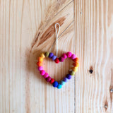 A colorful felt mini pompom love wreath ornament, shaped like a heart, rests on a warm-toned wooden surface. This handmade felt ornament is intricately crafted from numerous small, vibrant wool felt pompoms in red, pink, orange, yellow, green, blue, and purple, with a natural jute thread for hanging.