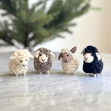 A row of unique wool felt mini sheep ornaments rests on a light marble surface, with a hint of green foliage in the soft background. These whimsical sheep feature woolly textures in cream, brown, white with brown ears, and black. Each has a roundish shape, small ears, small legs, and a simple stitched smiling face.