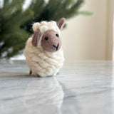 A charming mini wool felt sheep Christmas ornament rests on a reflective marble surface, with a blurred green backdrop. This needle felted sheep features a round, fluffy white body with a light brown face and ears, tiny black stitched eyes and mouth, and a scarf.