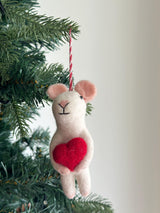 A charming wool felt mouse holding heart ornament is nestled among the green branches of a Christmas tree. This white mouse features light pink ears, tiny black eyes, and a simple black Y-shaped face. It clutches a bright red heart, with a red and white string attached for hanging.