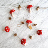 A whimsical felt mushroom and acorn garland rests on a white marble surface. This charming felt wool garland features alternating red mushrooms with white spots and tan acorns, strung along a thin twine. Its gentle curve creates an inviting display.