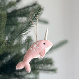 A charming pink needle felted narwhal ornament, made from 100% wool, hangs suspended from a vibrant green Christmas tree branch. This handmade, 3D felt narwhal features a pink body adorned with white spots, a white horn, and a sweet black eye, complete with a jute string for hanging.