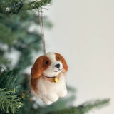 A handcrafted Cavalier King Charles Spaniel dog felt ornament is nestled among the green branches of a Christmas tree. This plush, textured ornament features a white and brown coat, a black string collar with a golden bell, and hangs by a brown jute string, ready to bring festive cheer.