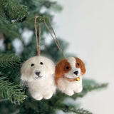 Two handmade felt Cavalier King Charles Spaniel dog ornaments hang nestled among the green needles of a Christmas tree. One is white, and the other is brown and white with a small golden bell, both featuring sweet expressions with black eyes and stitched mouths. Each needle felted dog is suspended by a rustic jute string.
