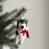 A wool felt Christmas husky ornament with a red scarf hangs gracefully from a Christmas tree branch. This charming handmade felt ornament is crafted primarily in white, black, and gray hues, accented by its vibrant red scarf, and suspended by a delicate white string.