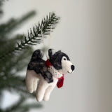A handcrafted felt husky Christmas ornament hangs from a Christmas tree branch, bathed in soft, diffused light. The fluffy, textured husky felt ornament features distinct gray and white markings. It wears a knitted red scarf and has a delicate cotton string attached for hanging, evoking a warm holiday mood.