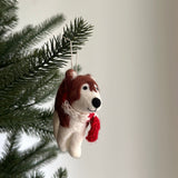 A handcrafted Christmas husky felt ornament with a red scarf hangs from a festive Christmas tree branch against a bright, plain background. The charming white and brown husky features a black stitched nose and visible felt textures. A cotton string is attached at the top for easy hanging, adding a touch of warmth and nostalgia to holiday decor.