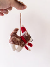 A playful Christmas husky felt ornament, crafted from brown and white felt, is held in a hand against a white background. This charming handmade ornament features a red and white Santa hat and a matching red scarf. A jute string extends from the top, ready for cheerful display.