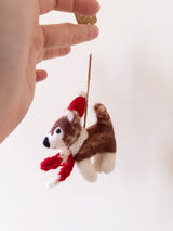 A female hand delicately holds a charming handcrafted Christmas husky felt ornament against a light background. This delightful felt husky ornament features brown and white fur, accented with a festive red Santa hat and a matching red scarf. It is suspended by a jute string.
