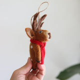 A cute Christmas reindeer felt ornament is gently held by a female hand. The brown reindeer features charming brown antlers, subtle white spots on its body, and wears a festive red scarf. A jute string is attached at the top, completing its look as a Christmas tree ornament.