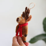 A cute Christmas reindeer felt ornament is held by a female hand against a bright background, creating a cheerful, festive scene. This handcrafted felt ornament is primarily brown with darker brown antlers and black hooves. A red scarf adorns its neck, and a jute string loop extends from its top for easy hanging.
