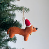 A vibrant felt Dachshund ornament hangs gracefully from a Christmas tree branch. This handmade piece features four adorable Dachshunds in cream, khaki, brown, and black, encircling a playful felt bone at its center. The charming scene is presented on a white felt frame, ready to hang with its jute string.