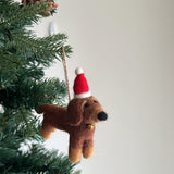 A single brown felt dachshund ornament hangs from a snow-dusted green Christmas tree branch, set against a plain white wall. This festive felt dachshund ornament features darker brown details, a black nose, and dark eyes, complete with a brown jute string for hanging.