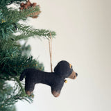 A playful felt dachshund dogs ornament hangs on a Christmas tree, secured by a jute string in a festive setting. This unique Christmas ornament features four needle-felted dachshunds in cream, khaki, brown, and black, each with characteristic short legs and long bodies. They are arranged on a white felt frame with a small felt bone at the center, adding a whimsical touch.