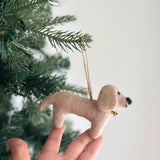 Wool felt dachshund ornaments are delicately presented by a female hand against a blurred Christmas tree, setting a cozy and festive ambiance. This charming ornament features four dachshunds in cream, khaki, brown, and black, each with floppy ears and stubby legs, surrounding a central felt bone. The playful arrangement is set on a white felt frame with a jute string for hanging.