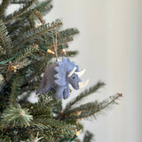 A light gray felt dinosaur triceratops ornament hangs on a festive green Christmas tree adorned with tiny light bulbs. This hand-stitched wool felt triceratops features a light gray body, a textured frill, and white horns, showcasing detailed craftsmanship. A jute string is attached to the top for easy hanging.