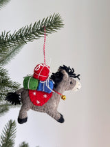 A wool felt donkey ornament with gifts hangs from a Christmas tree branch. The gray donkey features black hooves and mane, carrying stacked red, green, and blue gift packages tied with white strings. A red felt saddle with white snowflake designs and a golden bell adorn its neck, suspended by a red and white cotton string.