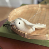 A pure white wool felt dove ornament with an olive twig in its beak is nestled inside a round, light brown woven basket. The delicate dove features a small black eye and green leaves on the twig, with a silver string attached for hanging. The basket rests on muted green and reddish-brown surfaces, creating a serene display.