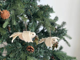 A white wool felt dove ornament with a green olive twig hangs on a green Christmas tree branch, visible against a plain white wall. The felt dove ornament is crafted from soft, pure white wool, holding a small green felt olive twig in its beak. A jute string is attached at the top for hanging, surrounded by lush green needles and a pine cone on the branch.