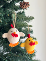 Two cheerful wool felt Easter chick ornaments hang on a green Christmas tree. These egg-shaped handmade felt ornaments are crafted from soft wool felt. One chick is cream, the other yellow, both featuring red wattles, orange beaks, and orange feet. Each handcrafted felt ornament has a sturdy hemp string for hanging, adding a charming, festive touch.