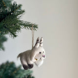 A handcrafted felt goat ornament hangs from a vibrant green Christmas tree branch, creating a festive and joyful atmosphere. This white needle-felted goat, made from organic wool, features dark gray markings on its feet and ears, a small pink nose, and a tiny gold bell around its neck. A jute string for hanging highlights its soft, handmade quality.