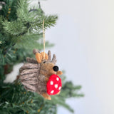 A charming wool felt hedgehog ornament hangs from a Christmas tree branch, suspended by a jute string against a soft white backdrop. The handcrafted hedgehog features a textured body, brown ears and limbs, and a black nose. It playfully holds a red mushroom with white spots, adding a whimsical touch to the Christmas tree.