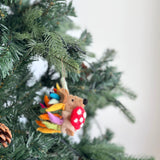 A playful wool felt hedgehog ornament hangs on a Christmas tree, evoking a festive mood. This unique Christmas ornament features a light brown hedgehog with a black nose, holding a red mushroom with white dots. Its colorful quills are yellow, green, and blue, with a brown jute string at the top for hanging. A pine cone sits in the lower-left corner, adding a natural touch.